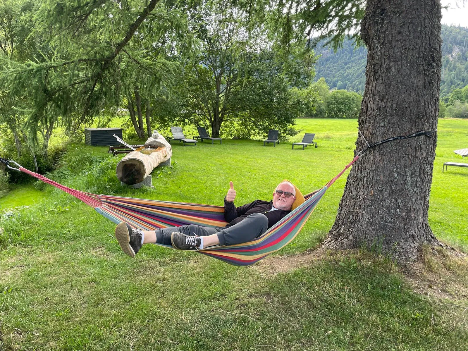 Ein Gast liegt im Garten in einer bunten Hängematte unter einem großen Baum und hebt den Daumen