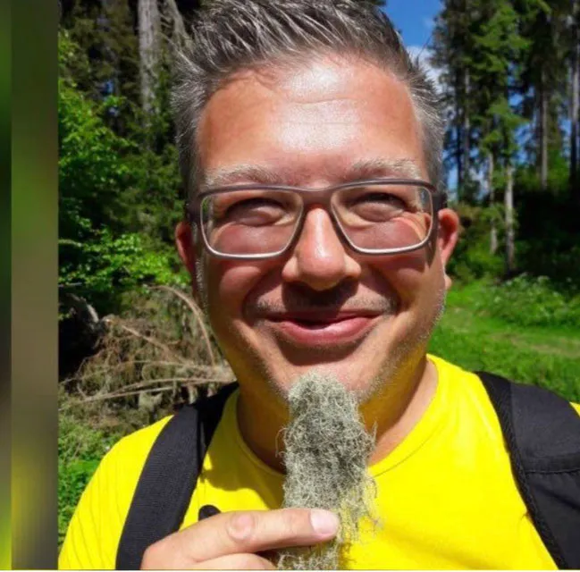 Marco mit Brille lächelt in die Kamera und hält eine weiße Pflanze in der Hand