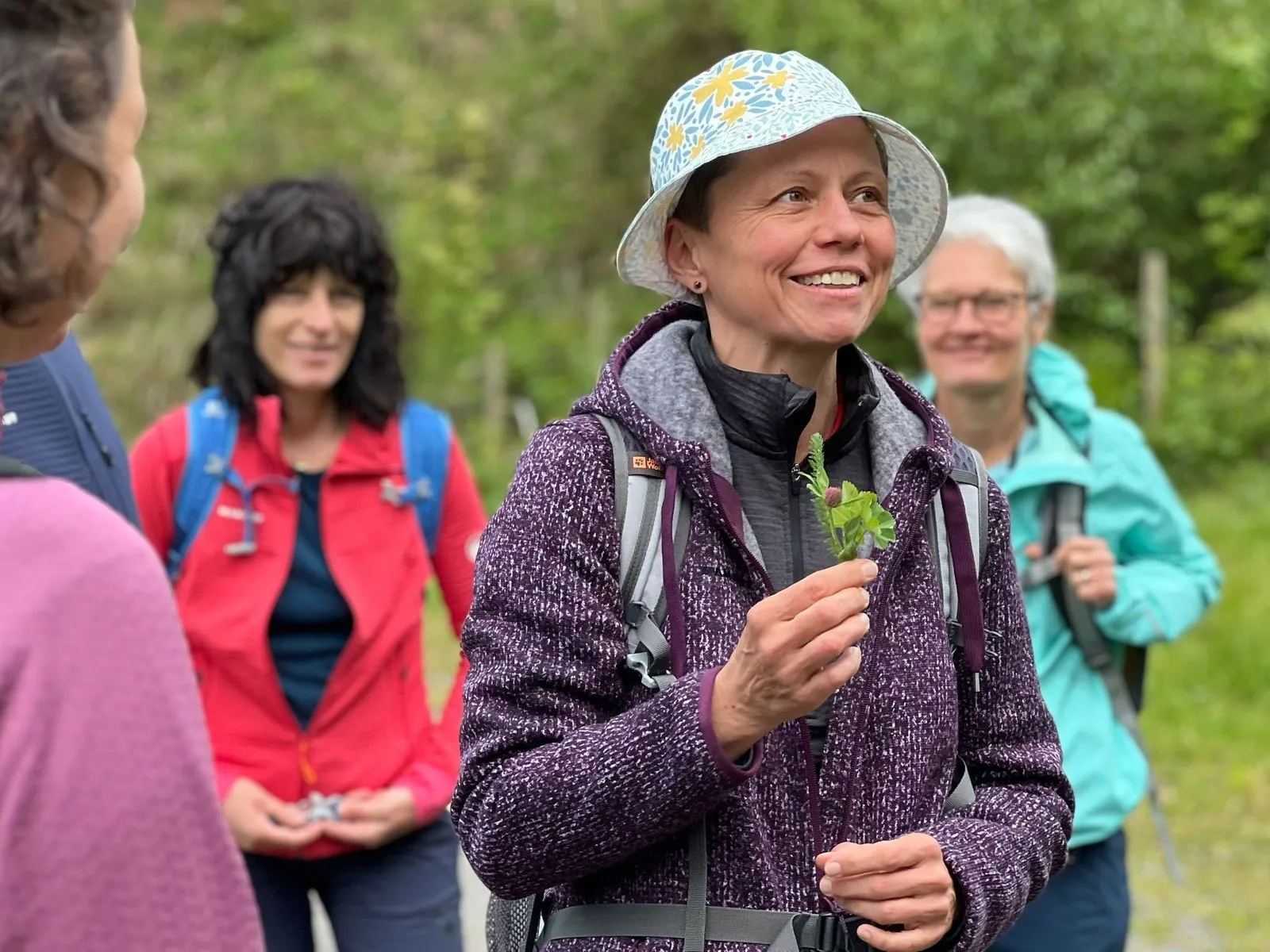 Unterwegs im Wald: Eine Frau mit Sonnenhut hält lächelnd eine Pflanze in der Hand, zwei Wanderinnen schauen zu
